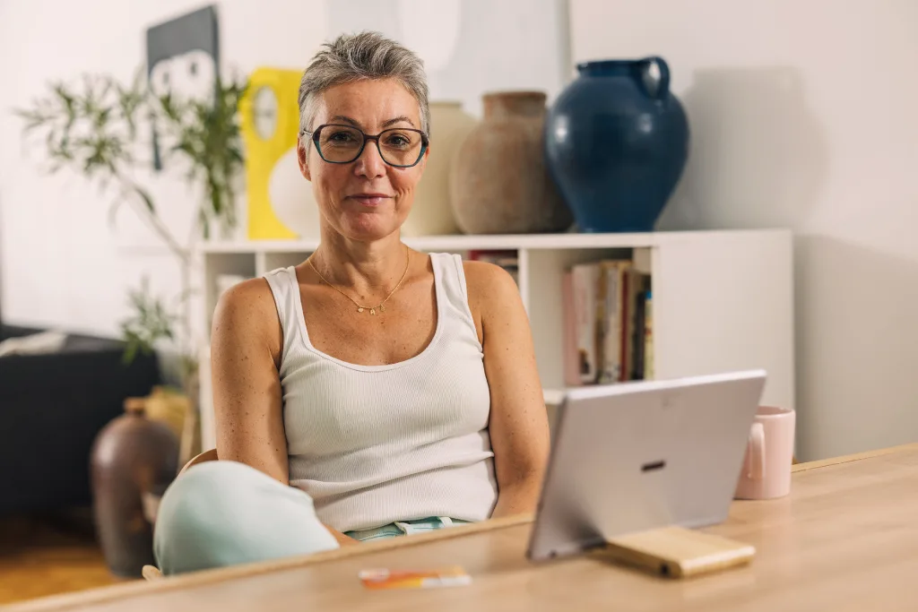 A woman with short grey hair and glasses sitting at a desk with a tablet, representing a virtual menopause consultation.