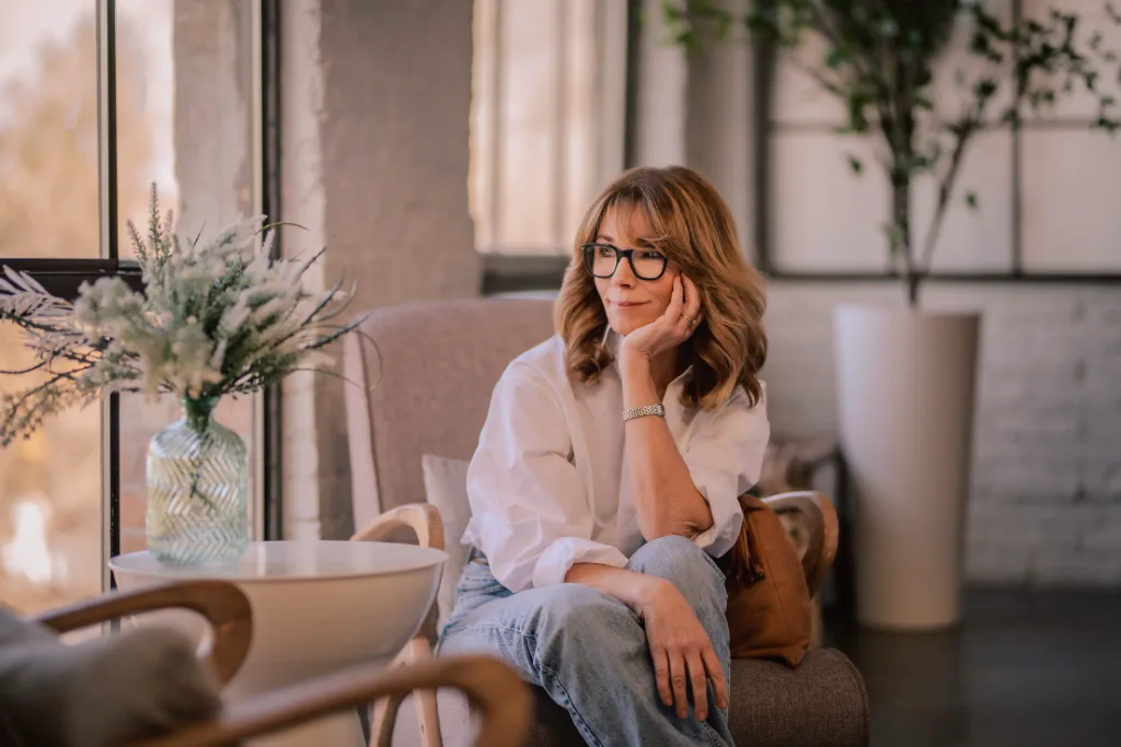 A thoughtful woman with glasses and a white shirt sitting in a modern, sunlit living room with plants.