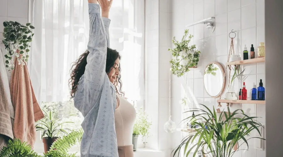 A woman stretching in a bright, sunlit bathroom filled with plants, illustrating healthy morning routines and increased energy levels for metabolic health in midlife.