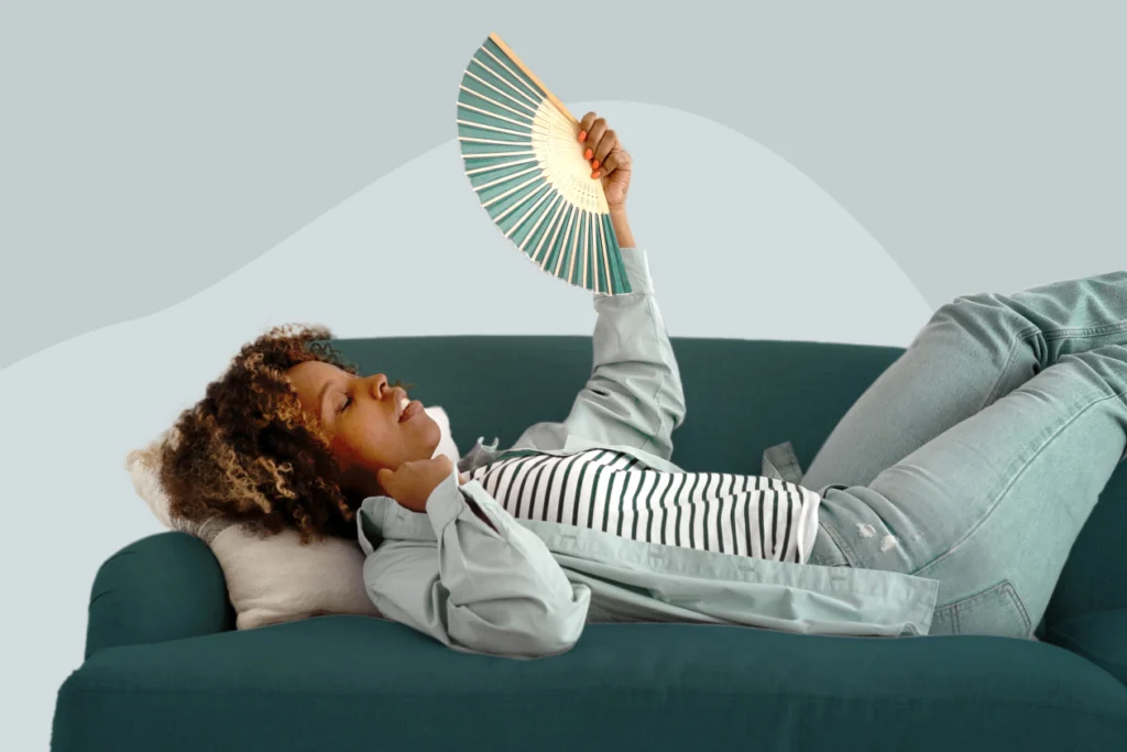 A woman lying on a green couch using a hand fan to cool down, illustrating menopause temperature dysregulation and hot flash symptoms.