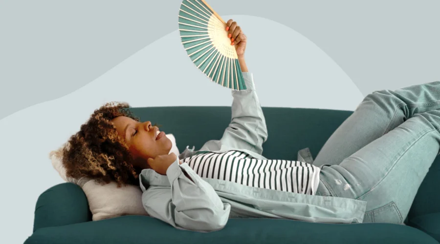A woman lying on a green couch using a hand fan to cool down, illustrating menopause temperature dysregulation and hot flash symptoms.