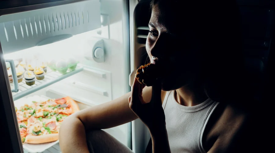 A woman eating a snack in the dark while illuminated by an open refrigerator, illustrating late-night cravings driven by perimenopause hormonal shifts.