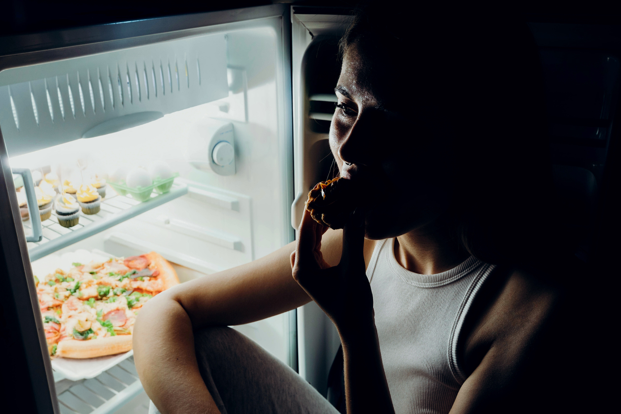 A woman eating a snack in the dark while illuminated by an open refrigerator, illustrating late-night cravings driven by perimenopause hormonal shifts.