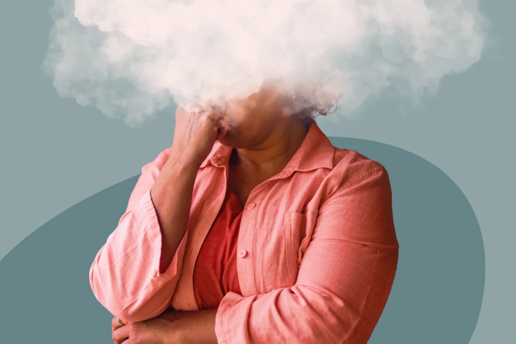 Femme en chemise corail, la main sous le menton et la tête cachée dans un nuage blanc, illustrant le brouillard mental à la ménopause