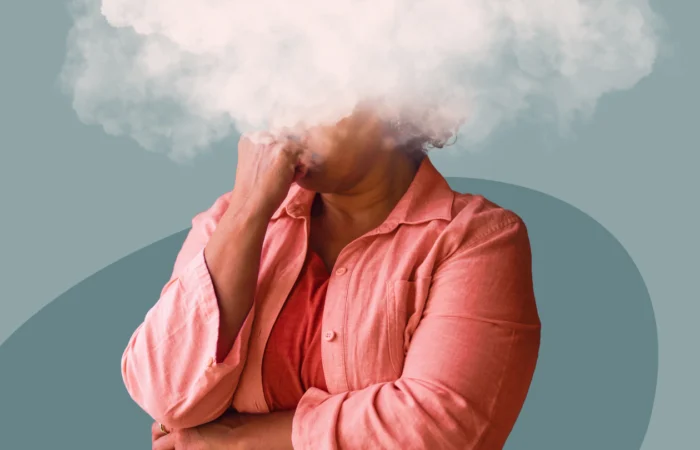 Blog-cover_brainfog Femme en chemise corail, la main sous le menton et la tête cachée dans un nuage blanc, illustrant le brouillard mental à la ménopause