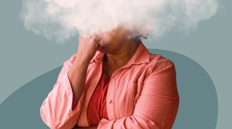 Femme en chemise corail, la main sous le menton et la tête cachée dans un nuage blanc, illustrant le brouillard mental à la ménopause